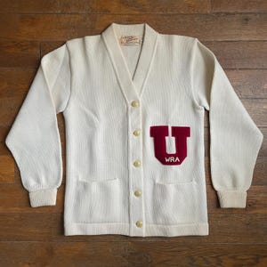 May include: White cardigan sweater with a red felt letter "U" and the text "WRA" on the left side. The sweater has a V-neck, long sleeves, and five buttons down the front.