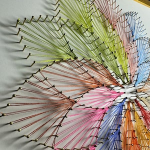 May include: A colourful string art piece with a circular design. The strings are arranged in a radial pattern, creating a starburst effect. The colours of the strings include red, orange, yellow, green, blue, and purple.