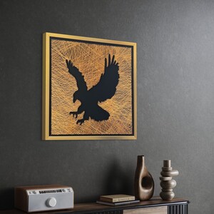 May include: A black eagle silhouette created with gold thread on a black canvas, framed in gold. The artwork is hanging on a dark gray wall above a wooden table with a vintage radio, a vase, and a stack of decorative stones.