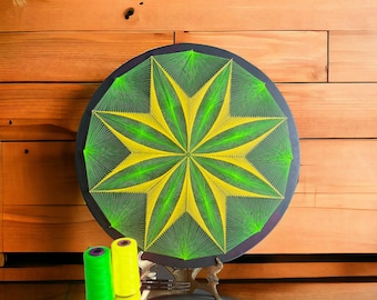 May include: A circular string art piece with a green and yellow starburst design. The art is mounted on a black circular frame and is displayed on a wooden surface. Two spools of thread, one green and one yellow, are in the foreground.