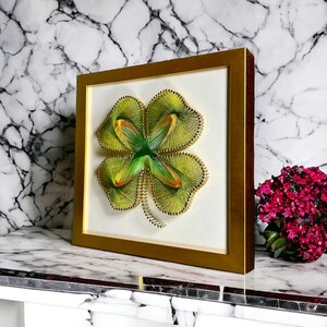 May include: A framed string art piece depicting a four-leaf clover in shades of green and yellow. The clover is surrounded by a white background and a gold frame.