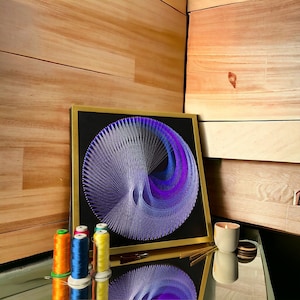 May include: A framed string art piece with a purple and blue swirl design on a black background. The frame is gold and there are spools of colourful thread in front of the frame.