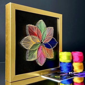 Könnte beinhalten: Ein farbenfrohes String-Art-Blumenmotiv in einem goldenen Rahmen. Die Blume besteht aus verschiedenen farbigen Fäden, die in einem kreisförmigen Muster um Nägel gewickelt sind. Der Hintergrund ist schwarz.