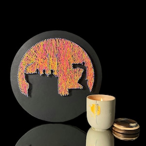 May include: A circular piece of art with a colorful string art design of a stylized elephant. A gray candle with the text "mundane" in gold lettering is in the foreground.