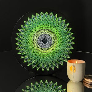 Op de afbeelding: Een groen en wit string art ontwerp in de vorm van een stervorm op een zwarte achtergrond. Het ontwerp is gemaakt met een verscheidenheid aan groene en witte draden, en het midden van de stervorm is een witte cirkel met een zwart centrum.