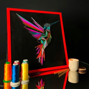 Könnte beinhalten: Ein lebendiges String-Art-Stück mit einem Kolibri. Das Kunstwerk hat einen schwarzen Hintergrund mit einem roten Rahmen. Der Kolibri ist mit farbigen Fäden gefertigt, darunter Pink, Grün, Blau und Orange. Garnrollen und eine brennende Kerze befinden sich im Vordergrund.