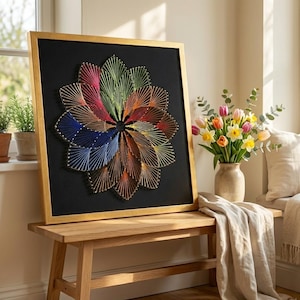 May include: A framed string art piece featuring a colorful floral design. The artwork is composed of various colored threads stretched across a black background, framed in gold. The piece is displayed on a wooden bench.