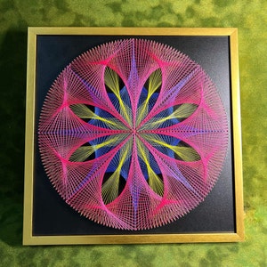 Puede incluir: Una pieza de arte de cuerda circular enmarcada con un diseño geométrico rosa, amarillo y azul sobre un fondo negro.
