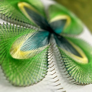May include: A green, yellow and blue string art piece in the shape of a four-leaf clover. The string is wrapped around nails hammered into a white surface.