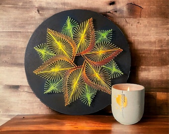 May include: A circular piece of art with a colorful string art design of a flower with yellow, orange, and green string on a black background. A gray candle with the text "Brewed Purpose" is in the foreground.