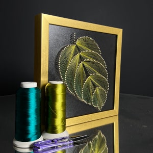 Könnte beinhalten: Ein goldfarbener Bilderrahmen mit schwarzem Hintergrund und einem grünen und gelben String-Art-Design eines Blattes. Im Vordergrund befinden sich zwei Garnrollen, eine grüne und eine türkisfarbene.