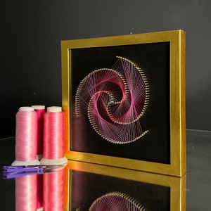 May include: A framed pink and purple string art piece in the shape of a spiral, with a black background. Three spools of pink thread are in the foreground.