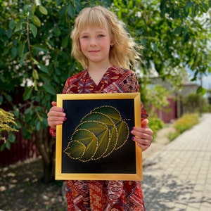 May include: A young person holding a framed piece of string art. The art depicts a green leaf with yellow string wrapped around nails. The frame is gold.