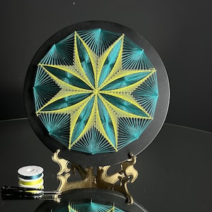 May include: A circular string art piece with a yellow and teal starburst design. The art is mounted on a black circular base and is displayed on a gold metal stand.