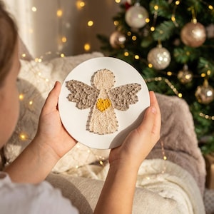 May include: A round, off-white decorative piece featuring a string art angel design. The angel has beige wings, a beige body, and a yellow heart. The piece is held in front of a Christmas tree with gold ornaments.