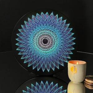 Puede incluir: Una pieza circular de arte de cuerda con un diseño geométrico azul, blanco y morado. El arte está montado en una tabla circular negra.