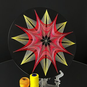 May include: A circular string art piece featuring a geometric star pattern created with red, pink, and yellow thread on a black board. The design radiates from a central point, with layered lines forming sharp, pointed star shapes.