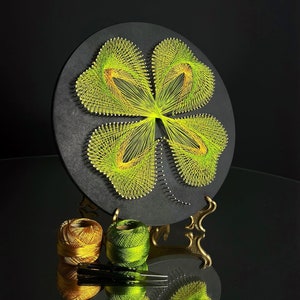 Puede incluir: Un tablero circular de madera negro con un diseño de trébol de cuatro hojas creado con hilo amarillo y verde. Dos carretes de hilo, uno dorado y uno verde, están en primer plano.