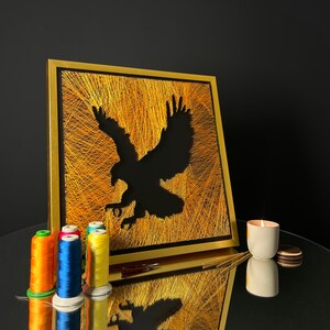 May include: A gold framed picture with a black silhouette of an eagle in flight. The eagle is surrounded by a background of yellow thread. The picture is sitting on a black surface with a lit candle and a small round object.