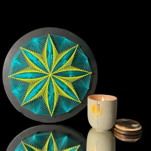May include: A teal and yellow string art starburst design on a black circular base. A lit candle in a gray ceramic container with a wooden lid sits next to the art piece. The candle has the text "aLaroma grapefruit".