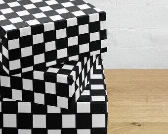 May include: Three stacked square boxes with a black and white checkerboard pattern. The boxes are on a wooden surface, with a white brick wall in the background. The boxes are different sizes, with the largest box at the bottom.