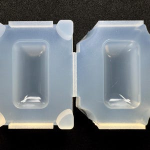 May include: A translucent, semi-flexible silicone mould featuring two rectangular cavities. The mould has a structured frame with four raised corner supports on each side, designed for casting rectangular shapes with a smooth, hollow interior finish.