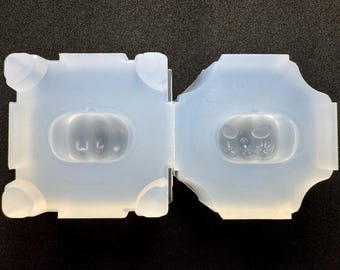 May include: A clear, flexible silicone mold for crafting. The mold is in two halves, each with a pumpkin shape. One pumpkin has a smiling face, the other a blank face. The mold has a square shape with rounded corners.