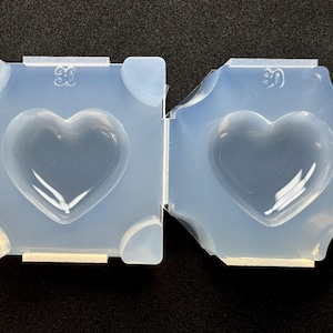 May include: Two translucent silicone moulds, each featuring a central heart-shaped cavity. The square-shaped mould on the left and the multi-faceted mould on the right both display the number 30 embossed near the top edge of their smooth surfaces.