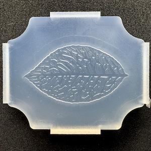 May include: A translucent, flexible silicone mold featuring a central embossed leaf design with intricate vein details. The mold has a rectangular shape with scalloped edges on all four sides, providing a clear impression surface for crafting.