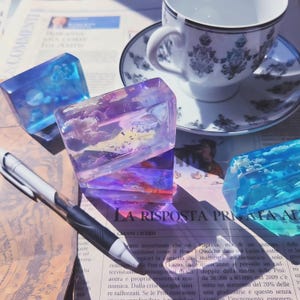 Op de afbeelding: Drie doorschijnende harsblokjes met wolken- en hemelontwerpen in blauwe, paarse en oranje tinten. Een witte en zwarte pen ligt op een krant met Italiaanse tekst. Een kop en schotel staan op de achtergrond.