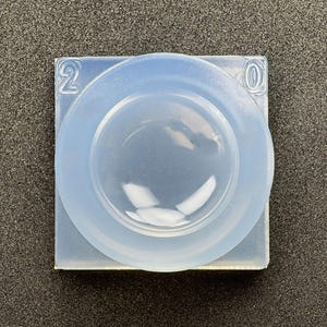 Peut inclure: Un moule en silicone bleu clair transparent, avec une base carrée et une forme circulaire bombée au centre. Les chiffres "2" et "0" sont en relief sur les coins supérieurs. Le moule est conçu pour le bricolage.
