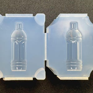 May include: A translucent silicone mold featuring two identical rectangular halves, each containing a recessed cavity shaped like a small water bottle with a narrow neck and a cylindrical body, designed for casting miniature bottle replicas.