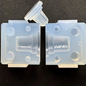 May include: A translucent silicone mould consisting of two rectangular halves and a separate top piece. The mould features a central cavity shaped like a stopper or plug, with circular alignment pegs and holes visible on the flat surfaces of the mould.
