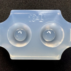 May include: A transparent silicone mould featuring two circular depressions. The rectangular mould has rounded edges and small tabs. The text "18-L" is imprinted on the top. Ideal for crafting.