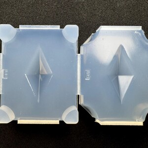 May include: Two-part, light blue silicone mold. Each part is square with rounded corners and a diamond-shaped indentation. The mold has a textured surface and is designed for crafting or casting.