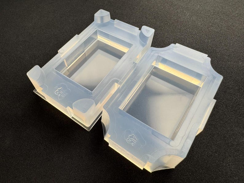 Op de afbeelding: Twee doorschijnende siliconen mallen met rechthoekige holtes en decoratieve randen. Elke mal heeft een verhoogde hoek en een klein reli&euml;fontwerp. De mallen zijn ontworpen voor knutselen en staan tegen een donkere achtergrond.