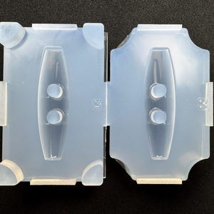 May include: A clear, two-part silicone mold for crafting. Each half features a central, elongated cavity with two circular indentations. The mold is designed with tabs and a connecting feature for alignment. The number "50" is visible on the mold.