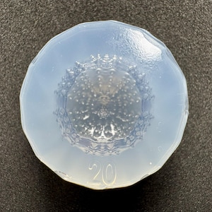 Pode incluir: Um molde de silicone circular e translúcido com uma borda facetada e um padrão interior detalhado e texturizado que se assemelha a um ouriço-do-mar. O número 20 está claramente gravado na borda inferior do molde, que tem um acabamento liso e brilhante.