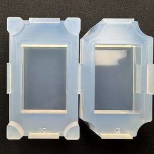 Könnte beinhalten: Zwei hellblaue Silikonformen, rechteckig, mit einem zentralen, vertieften Bereich. Eine Form hat abgerundete Ecken, während die andere ein dekoratives, geschwungenes Design aufweist. Beide haben kleine Laschen und sind wahrscheinlich zum Basteln oder Gießen gedacht.