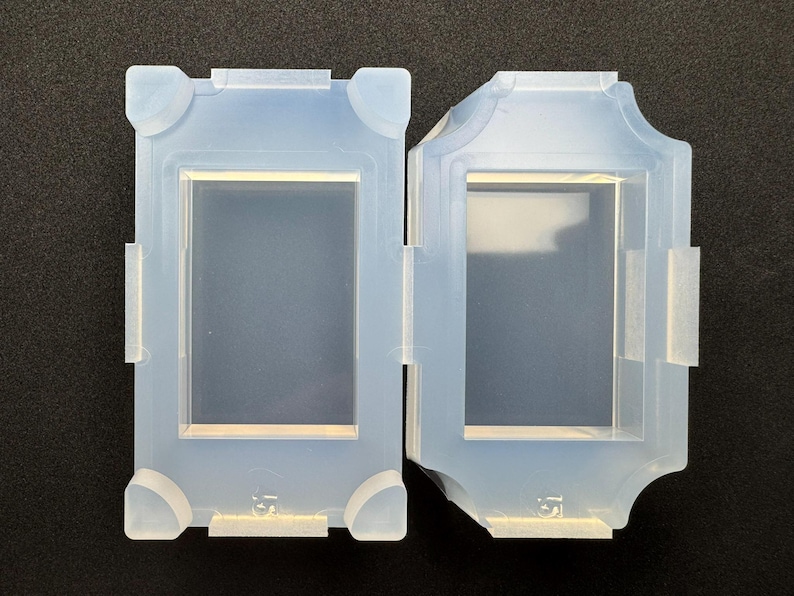 Op de afbeelding: Twee lichtblauwe siliconen mallen, rechthoekig van vorm, met een centraal verzonken gebied. E&eacute;n mal heeft afgeronde hoeken, terwijl de andere een decoratief, gebogen ontwerp heeft. Beide hebben kleine lipjes en zijn waarschijnlijk bedoeld voor knutselen of gieten.