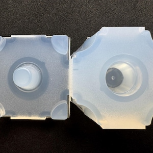 May include: Two translucent, light blue silicone molds. One is square with rounded corners and a central cylinder. The other is a six-sided shape with a central hole. Both have a matte finish.