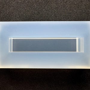 以下が含まれることがあります： 長方形の半透明シリコンモールドで、長方形のくぼみがあります。モールドの側面には小さなタブが付いています。暗い表面にオブジェクトがあります。