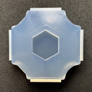 Può includere: Uno stampo in silicone traslucido e semi-flessibile caratterizzato da una forma a croce a quattro lati con bordi arrotondati e un'indentazione esagonale centrale. Lo stampo ha una finitura liscia e opaca, ideale per colate o artigianato.