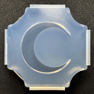 Könnte beinhalten: Eine durchscheinende, halbflexible Silikonform mit einer zentralen Mondsichelform. Die Form hat einen ausgeprägten achteckigen Außenrand mit vier flachen, geraden Kanten und vier abgerundeten Ecken, die zum Gießen von Harz oder anderen Materialien dient.