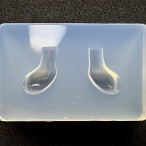 May include: A rectangular, translucent silicone mold featuring two identical, hollowed-out cavities shaped like small, upright socks or booties. The mold has a smooth, matte finish and is designed for casting small objects in a mirrored, side-by-side orientation.
