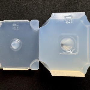 May include: Two translucent, square-shaped molds with rounded corners. Each mold has a central, circular indentation and the number "15" embossed on the surface. The molds are made of a flexible, clear material.
