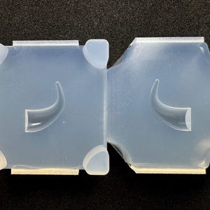 May include: Two translucent, light blue silicone molds. Each mold is square with rounded corners and a curved horn shape in the center. The molds have small corner supports and a rectangular base.
