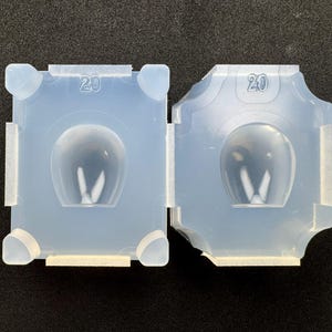 Puede incluir: Dos moldes de silicona azul claro, cada uno con una cavidad redondeada y una forma de 'V' en el interior. Los moldes están numerados con el número '20' y tienen pestañas y soportes en las esquinas. Diseñado para manualidades o fundición.