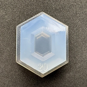 May include: A clear, hexagonal mould with a central geometric design and the number "20" at the bottom. The mould is made of a translucent material, and the edges are slightly textured. The background is a dark grey.