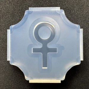 Könnte beinhalten: Eine durchscheinende, halbflexible Silikonform mit einem mittigen Ausschnitt in Form eines weiblichen Geschlechtssymbols. Die Form ist quadratisch mit abgerundeten, gewellten Kanten und flachen Laschen an allen vier Seiten.